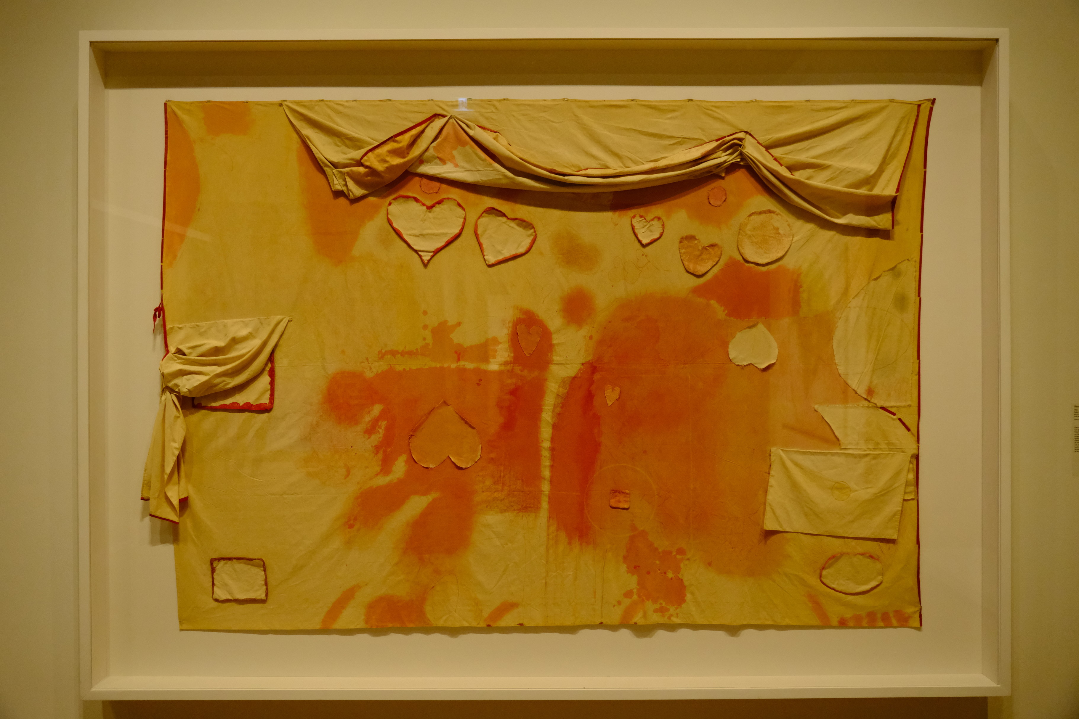A piece of textile art displayed within a large, thick white frame features a large, rectangular yellow fabric with various orange and reddish-orange patches throughout. Several fabric cutouts in the shape of hearts are attached to the main fabric. Draped and folded pieces of the same yellowish fabric hang from the top edge and along the left side of the central piece. The entire framed work is set against a plain, neutral-colored background